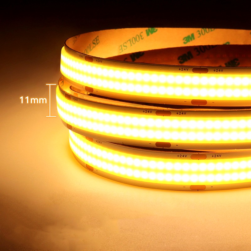 Close-up of 11mm wide 24V COB LED strip lights emitting warm yellow light, coiled with visible 3M adhesive backing, ideal for ambient lighting applications.