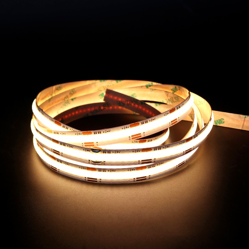 Coiled 24V COB LED strip light glowing warm white, placed on a dark surface with 3M adhesive backing visible.