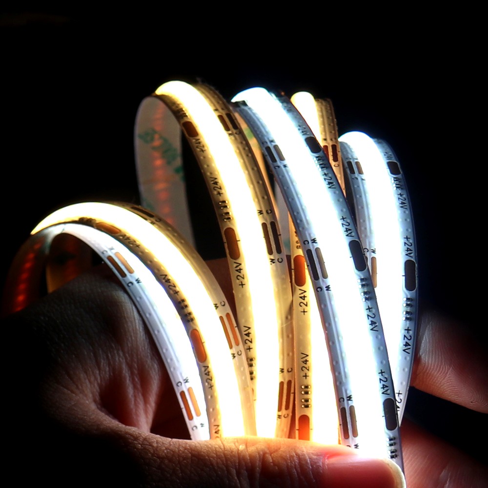A hand holding multiple 24V COB LED strip lights, each emitting different color temperatures from warm to cool white, demonstrating brightness levels and flexibility.