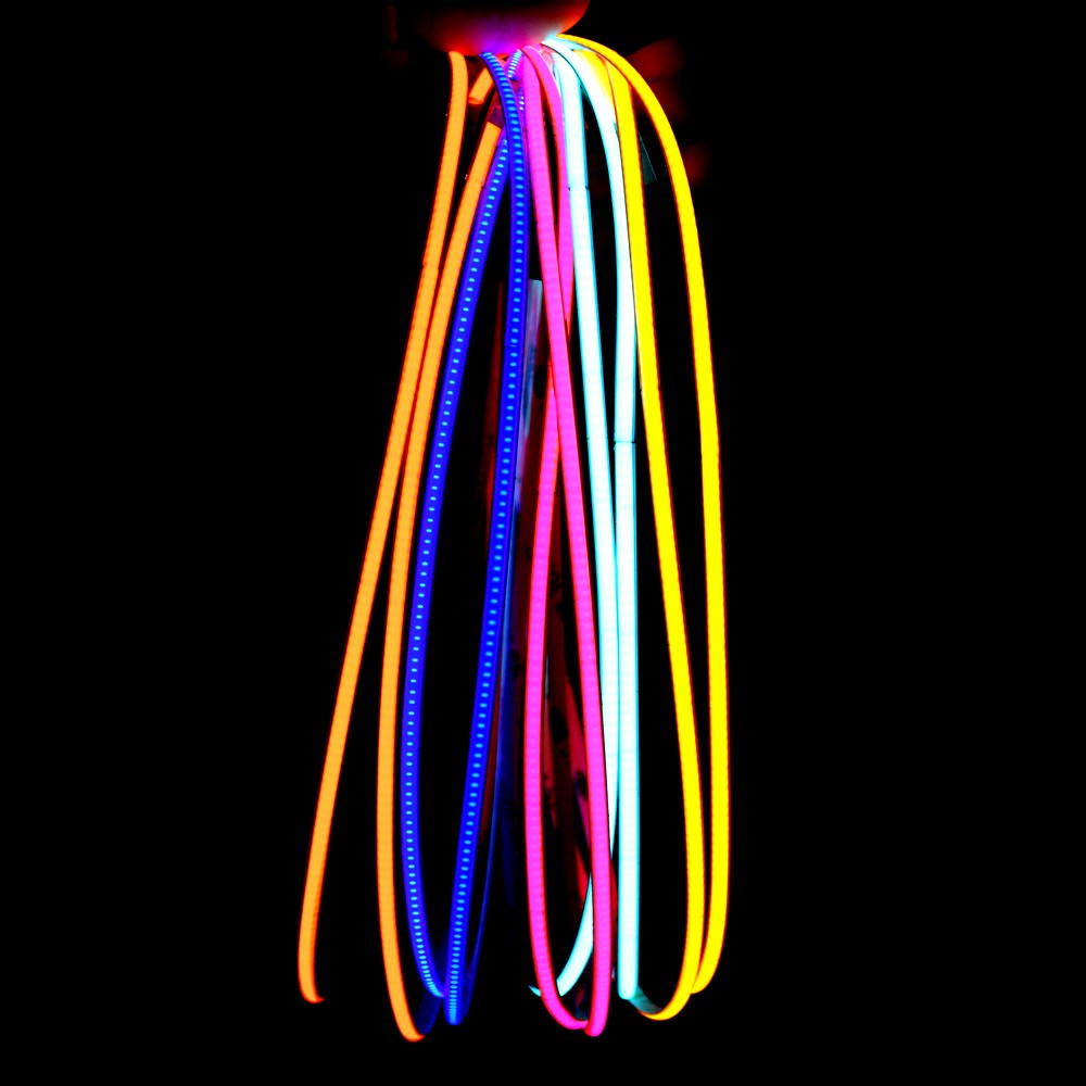 Various colors of neon flex LED strip lights including orange, blue, pink, and cyan, glowing brightly in a dark background and hanging vertically for display.