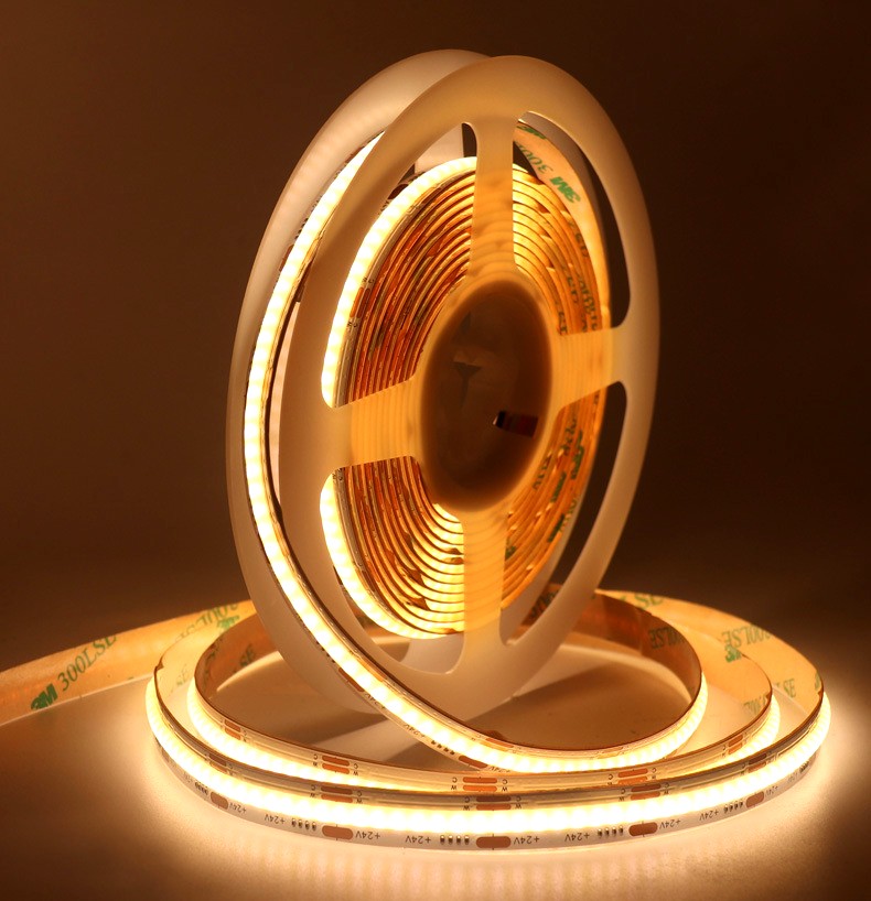 Warm white 24V COB LED strip light rolled on a plastic reel, glowing brightly with uniform illumination and visible 3M 300LSE adhesive backing for easy installation.